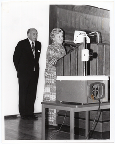B&W photo of a woman standing at a tall wooden podium. There is a man standing behind her. Beside the podium is a large slide projector on a table.