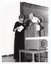 B&W photo of a woman standing at a podium on a stage. She is looking at the contents of an envelope in her hand, while a man standing beside her looks on. On a small table, beside the podium, is a large slide projector.