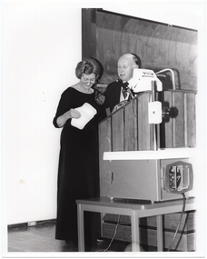 B&W photo of a woman standing at a podium on a stage. She is looking at the contents of an envelope in her hand, while a man standing beside her looks on. On a small table, beside the podium, is a large slide projector.
