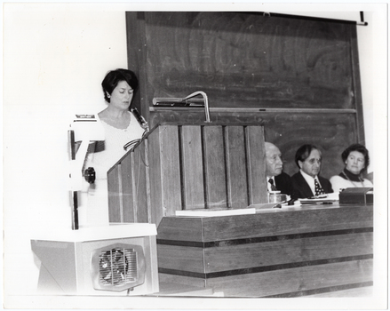 B&W photo of a woman standing at a podium built into a large wooden desk. Three people are seated behind the desk. On a small table, beside the podium, is a large slide projector.