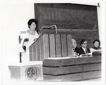 B&W photo of a woman standing at a podium built into a large wooden desk. Three people are seated behind the desk. On a small table, beside the podium, is a large slide projector.