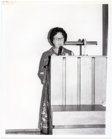 B&W photo of a woman standing at a large wooden podium, speaking.