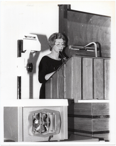 B&W photo of a woman standing at a large wooden podium, speaking.