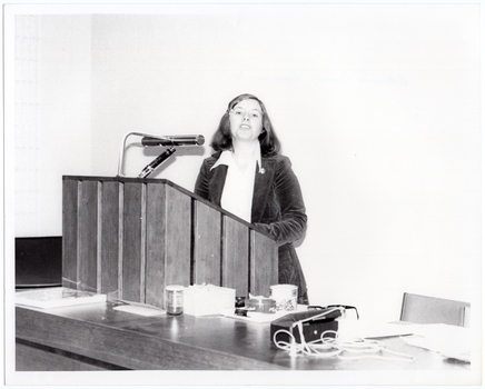 B&W photo of a woman standing at a podium, speaking. The podium is built into a large wooden desk.