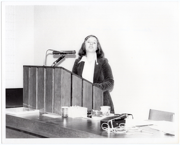 B&W photo of a woman standing at a podium, speaking. The podium is built into a large wooden desk.