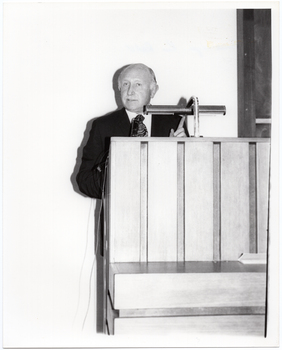B&W photo of a man standing at a podium built into a large wooden desk.