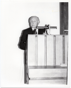B&W photo of a man standing at a podium built into a large wooden desk.