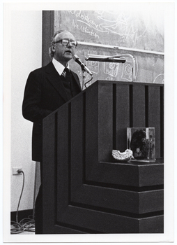 B&W photo of a man standing at a podium built into a large wooden desk. Behind him is a large blackboard with chalk markings.