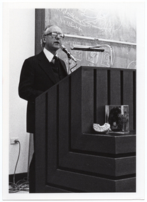 B&W photo of a man standing at a podium built into a large wooden desk. Behind him is a large blackboard with chalk markings.
