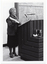 B&W photo of a woman standing at a podium built into a large wooden desk. She is holding up a rectangular framed trophy. Behind her is a large blackboard with chalk markings.