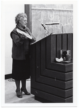 B&W photo of a woman standing at a podium built into a large wooden desk. She is holding up a rectangular framed trophy. Behind her is a large blackboard with chalk markings.