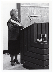 B&W photo of a woman standing at a podium built into a large wooden desk. She is holding up a rectangular framed trophy. Behind her is a large blackboard with chalk markings.