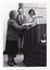 B&W photo of three people standing behind a podium built into a large wooden desk. A woman is passing a rectangular framed trophy to another woman. Standing in between and slightly behind them is a man, smiling. Behind them is a large blackboard with chalk markings.