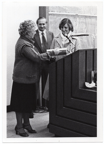 B&W photo of three people standing behind a podium built into a large wooden desk. A woman is passing a rectangular framed trophy to another woman. Standing in between and slightly behind them is a man, smiling. Behind them is a large blackboard with chalk markings.
