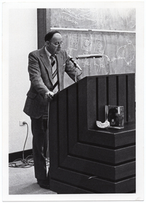 B&W photo of a man standing at a podium built into a large wooden desk. Behind him is a large blackboard with chalk markings.