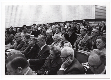 B&W photo of a large group of people seated on rows of seats in a large hall.