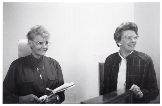 B&W photo of two women standing behind a podium (partially obscured). One of the women is holding an award trophy.