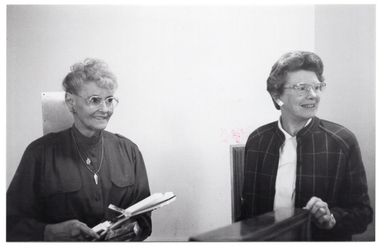 B&W photo of two women standing behind a podium (partially obscured). One of the women is holding an award trophy.