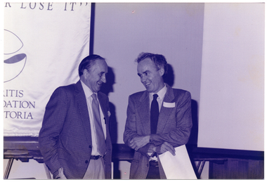 B&W photo of two men standing in front of a long trestle table. They are laughing. The trestle table is positioned against the wall. Beside the table is a large vertical banner containing the text, "Move It Or Lose It" (partially obscured and a logo (partially obscured).
