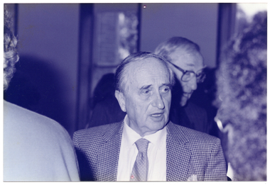 B&W close up photo of a man standing among a group of people, indoors. He appears to be speaking to someone.