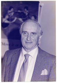 B&W informal portrait of a man standing indoors. His gaze is focused slightly to one side of the camera. In the background, there is a large "Move It Or Lose It" water exercise poster on the wall.