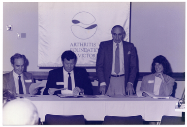 B&W photo of a man standing behind a long rectangular table at which three others are seated. He is looking straight ahead and speaking. Two of the three seated people are looking down at some paperwork, while the other is looking to one side, beyond the camera. The people seated are all smiling, to varying degrees. On the wall behind them is a large Arthritis Foundation of Victoria banner.