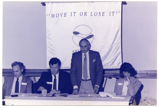 B&W photo of a man standing behind a long rectangular table at which three others are seated. He is speaking and looking down slightly. The people seated are all looking down at some paperwork on the table. On the wall behind them is a large banner containing the text, "MOVE IT OR LOSE IT", "ARTHRITIS FOUNDATION OF VICTORIA" (partially obscured), and a logo.