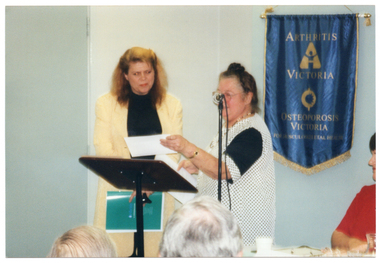 COL photo of two women standing at a lectern. One of the women is holding up an envelope in one hand while holding a piece of paper in her other hand. A third woman is seated at a table beside them. On the wall behind them is a blue banner with gold fringing and tassles. On the banner is the text, "ARTHRITIS VICTORIA" with a corresponding logo below, followed by the text "OSTEOPOROSIS VICTORIA" with a corresponding logo below that. At the bottom of the banner is the text, "FOR MUSCULOSKELETAL HEALTH". In the foreground, there is a (partially obscured) view of the back of two other people's heads.