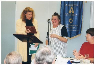 COL photo of two women standing at a lectern. One of the women is holding an award plaque in one hand, while holding a green folder with papers in her other hand. A third woman, applauding, is seated at a table beside them. On the wall behind them is a blue banner with gold fringing and tassles. On the banner is the text, "ARTHRITIS VICTORIA", with a corresponding logo below, followed by the text, "OSTEOPOROSIS VICTORIA", with a corresponding logo below that. At the bottom of the banner is the text, "FOR MUSCULOSKELETAL HEALTH". In the foreground, there is a (partially obscured) view of the back of the heads of two other people.