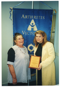 COL photo of two women posing for a photo, facing the camera. One of the women is holding up a framed award. On the wall behind them is a blue banner with gold text, fringing and tassles. On the banner is the text "ARTHRITIS VICTORIA" (partially obscured).