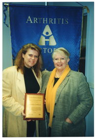 COL photo of two women posing for a photo, facing the camera. One of the women is holding up a framed award. On the wall behind them is a blue banner with gold text, fringing and tassles. On the banner is the text "ARTHRITIS VICTORIA" (partially obscured).