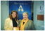 COL photo of two women posing for a photo, facing the camera. One of the women is holding up a framed award. On the wall behind them is a blue banner with text, gold fringing and tassles. On the banner is the text "ARTHRITIS VICTORIA" (partially obscured). On another wall is a clock, several light switches, and a small brown sign titled, "NOTICE TO ALL".