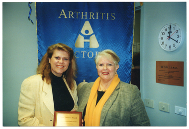 COL photo of two women posing for a photo, facing the camera. One of the women is holding up a framed award. On the wall behind them is a blue banner with text, gold fringing and tassles. On the banner is the text "ARTHRITIS VICTORIA" (partially obscured). On another wall is a clock, several light switches, and a small brown sign titled, "NOTICE TO ALL".