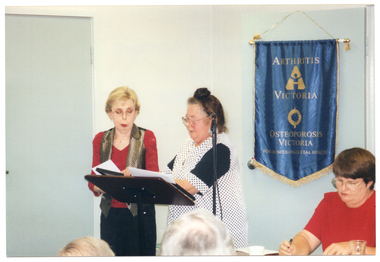COL photo of two women standing at a lectern. One of the women is presenting a framed award to the other woman, who is holding a white envelope. A third woman is seated at a table beside them - she is holding a pen in one hand. On the wall behind them is a blue banner with gold fringing and tassles. On the banner is the text "ARTHRITIS VICTORIA" with a corresponding logo below, followed by the text "OSTEOPOROSIS VICTORIA" with a corresponding logo below that. At the bottom of the banner is the text "FOR MUSCULOSKELETAL HEALTH". In the foreground, there is a (partially obscured) view of the back of the heads of two other people.