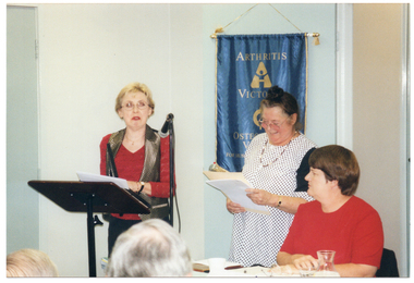 COL photo of a woman standing behind a lectern, on which sits a framed award and a white envelope. A second woman is standing beside her. She is looking down at a manila folder and some paperwork which she is holding in her hands. A third woman is seated at a table beside them. She is looking at the woman behind the lectern. On the wall behind them is a blue banner with gold fringing and tassles. On the banner is the text "ARTHRITIS VICTORIA" with a corresponding logo below, followed by the text "OSTEOPOROSIS VICTORIA" with a corresponding logo below that. At the bottom of the banner is the text "FOR MUSCULOSKELETAL HEALTH". In the foreground, there is a (partially obscured) view of the back of the heads of two other people.