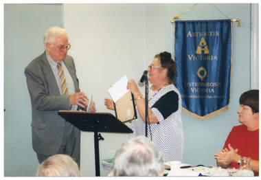 COL photo of a man and a woman standing behind a lectern. The man is holding a white envelope in one hand while his other hand is poised to receive a framed award from the woman. She also appears to be holding a white envelope in her hand, which is placed behind the framed award. A second woman is seated at a table beside them - she is applauding. On the wall behind them is a blue banner with gold fringing and tassles. On the banner is the text "ARTHRITIS VICTORIA" with a corresponding logo below, followed by the text "OSTEOPOROSIS VICTORIA" with a corresponding logo below that. At the bottom of the banner is the text "FOR MUSCULOSKELETAL HEALTH". In the foreground, there is a (partially obscured) view of the back of the heads of two other people.