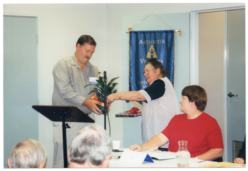 COL photo of a man and a woman standing behind a lectern. The woman is passing a potted plant (with an orange ribbon tied around the pot), to the man. In her other hand, she is holding a gift-wrapped book. A second woman, looking on, is seated at a table beside them. On the wall behind them is a blue banner with gold fringing and tassles. It contains the (partially obscured) text, "ARTHRITIS VICTORIA" with a corresponding logo below, followed by the text, "OSTEOPOROSIS VICTORIA", with a corresponding logo below that. At the bottom of the banner (obscured) is the text, "FOR MUSCULOSKELETAL HEALTH". In the foreground, there is a (partially obscured) view of the back of the heads of two other people.