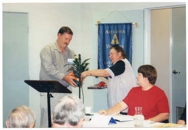 COL photo of a man and a woman standing behind a lectern. The woman is passing a potted plant (with an orange ribbon tied around the pot), to the man. In her other hand, she is holding a gift-wrapped book. A second woman, looking on, is seated at a table beside them. On the wall behind them is a blue banner with gold fringing and tassles. It contains the (partially obscured) text, "ARTHRITIS VICTORIA" with a corresponding logo below, followed by the text, "OSTEOPOROSIS VICTORIA", with a corresponding logo below that. At the bottom of the banner (obscured) is the text, "FOR MUSCULOSKELETAL HEALTH". In the foreground, there is a (partially obscured) view of the back of the heads of two other people.