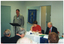 COL photo of a man standing behind a lectern, speaking. Seated at a table beside him are three people (one predominantly obscured). On the wall behind them is a blue banner with gold fringing and tassles. On the banner is the text "ARTHRITIS VICTORIA" with a corresponding logo below, followed by the text "OSTEOPOROSIS VICTORIA" with a corresponding logo below that. At the bottom of the banner is the text "FOR MUSCULOSKELETAL HEALTH". In the foreground, there is a (partially obscured) view of the back of the heads of four other people.