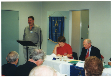 COL photo of a man standing behind a lectern, speaking. Seated at a table beside him are three people (one predominantly obscured). On the wall behind them is a blue banner with gold fringing and tassles. On the banner is the text "ARTHRITIS VICTORIA" with a corresponding logo below, followed by the text "OSTEOPOROSIS VICTORIA" with a corresponding logo below that. At the bottom of the banner is the text "FOR MUSCULOSKELETAL HEALTH". In the foreground, there is a (partially obscured) view of the back of the heads of four other people.