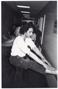 B&W photo of several people doing some stretching exercises in a long, narrow hallway. There is a handrail on one wall, and a brick wall without a handrail on the opposite wall. Some people have one leg up on the handrail while others have their leg placed up high on the brick wall opposite. Each person exercising is bending their torso towards the ankle of the raised leg. There are two people who appear to be standing while the others are doing the exercises.