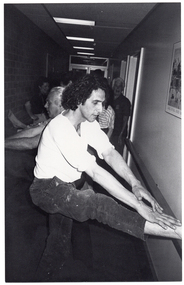 B&W photo of several people doing some stretching exercises in a long, narrow hallway. There is a handrail on one wall, and a brick wall without a handrail on the opposite wall. Some people have one leg up on the handrail while others have their leg placed up high on the brick wall opposite. Each person exercising is bending their torso towards the ankle of the raised leg. There are two people who appear to be standing while the others are doing the exercises.