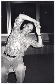 B&W photo of a man sitting on a chair. He has his hands clasped above his head while he leans his torso to one side. He is in a room with large windows along the wall behind him. It appears to be dark outside.