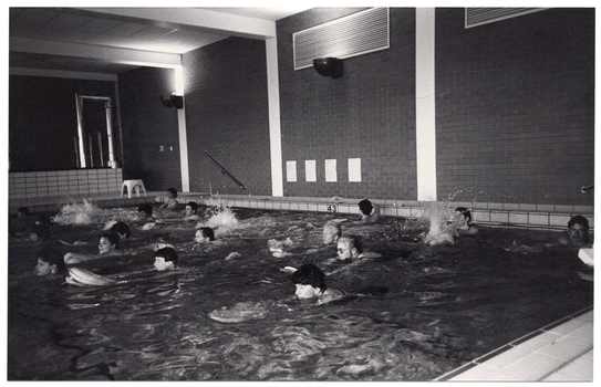 B&W photo of a group of people exercising in an indoor swimming pool. They are each holding a kickboard, with their arms outstretched, to propel themselves forward in the water.