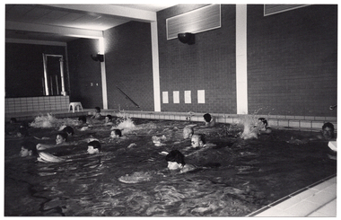 B&W photo of a group of people exercising in an indoor swimming pool. They are each holding a kickboard, with their arms outstretched, to propel themselves forward in the water.