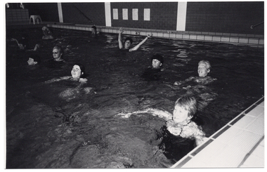 B&W photo of a group of people exercising in a swimming pool. A few of the people in the foreground are looking towards the camera. One woman has the back has her arms outstretched as she leans backwards in the water.