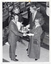 B&W photo of a man presenting a silver coffee service on a silver platter to a woman. Standing nearby, another woman and man look on. Beside and behind the group are several paintings arranged on various display boards. The photo is taken from a high angle, looking down toward the subjects.