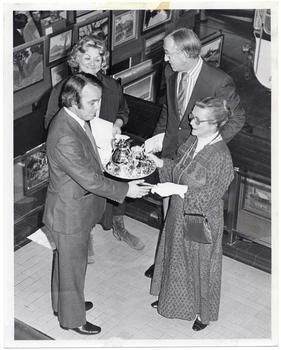 B&W photo of a man presenting a silver coffee service on a silver platter to a woman. Standing nearby, another woman and man look on. Beside and behind the group are several paintings arranged on various display boards. The photo is taken from a high angle, looking down toward the subjects.