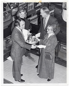 B&W photo of a man presenting a silver coffee service on a silver platter to a woman. Standing nearby, another woman and man look on. Beside and behind the group are several paintings arranged on various display boards. The photo is taken from a high angle, looking down toward the subjects.