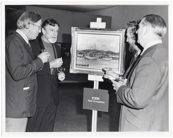 B&W photo of four men gathered around a painting sitting on an easel. One of the men is holding a beer glass, while the other three men are each holding a glass of champagne. Affixed to the easel (below the painting) is a sign with the text, "WINNER Open Competition".